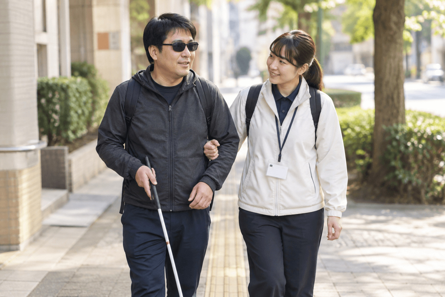 白杖を持つ利用者に支援者が寄り添い、腕を支えながら歩道を歩いている様子